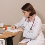 A female doctor with a stethoscope writing notes at a desk indoors.