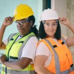 Two female construction workers wearing hard hats and safety gear, standing confidently indoors.